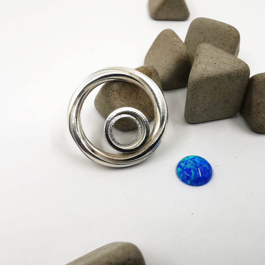 A bespoke pair of Twist Continuum earrings in sterling silver with gold plated detailing, featuring 8mm lab grown opal cabochon stones with vivid blue and green colour play. The stones are set within the lower section of the circular form, creating contrast between structured geometry and shifting colour. Created as a commission for Fire & Iron Gallery, this piece explores movement, balance and contemporary circular design.